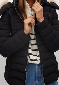 Zwarte poofy jas met een capuchon, voorzien van een ritssluiting en geblokte details. Draag over een witte trui met marineblauwe strepen.