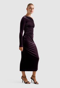 Violetā samta midi kleita ar garām piedurknēm un drapējuma detaļām, apvienota ar melnām siksnu kurpēm uz papēžiem. Gluda tekstūra un pieguļoša forma.