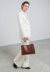 Braune Lederhandtasche mit strukturiertem Design und einem Griff oben, die ein minimalistisches Design aufweist. Das Modell trägt einen weißen maßgeschneiderten Anzug und schwarze High Heels.