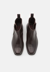 Braune Leder-Ankle-Boots mit einer glatten, strukturierten Oberfläche, quadratischer Zehenpartie, seitlichem Reißverschluss und kontrastierendem roten Tab-Detail an der Ferse.