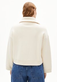 Cropped, cream-colored sweatshirt with a wide collar and ribbed cuffs, paired with blue tapered jeans featuring back pockets.