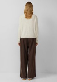 Beżowy sweter o fakturowanym wykończeniu i ściągaczu, zestawiony z brązowymi spodniami o szerokich nogawkach oraz butami z białą podeszwą. Przedstawiono widok od tyłu.