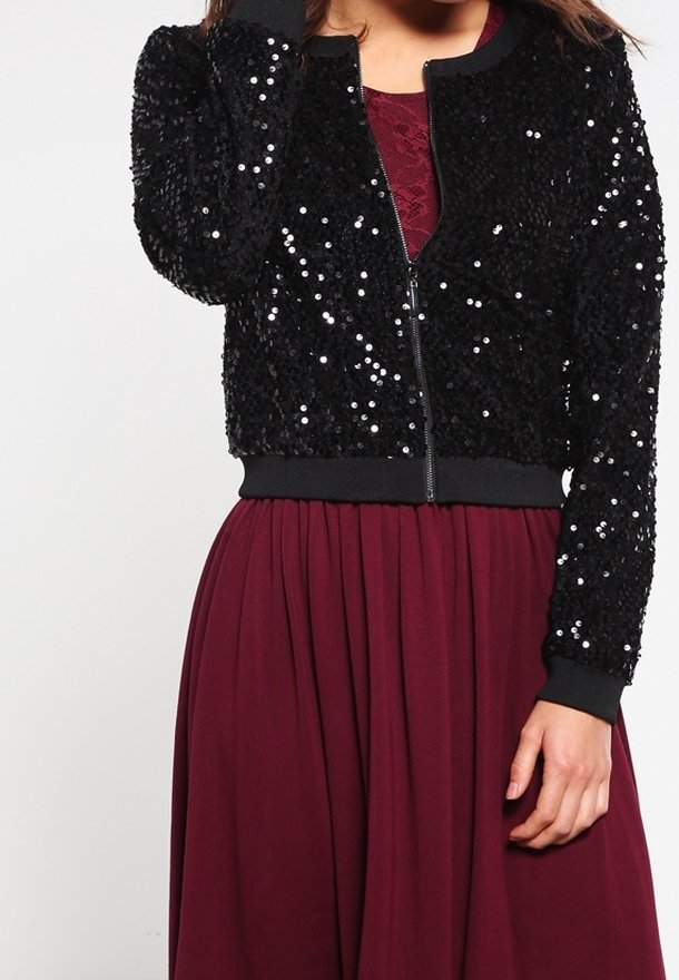 Personne portant une veste noire à sequins sur une robe bordeaux en dentelle et plissée, debout devant un fond clair uni.