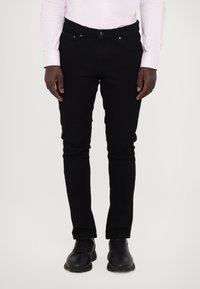 Jean skinny noir à texture lisse, design à cinq poches, bouton métallique et détails de couture subtils ; associé à des baskets noires.