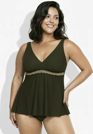 Uśmiechnięta kobieta z ciemnymi włosami sięgającymi ramion, ubraną w oliwkowo-zielony teksturowany tankini ze złotym wykończeniem, stojąca z jedną ręką na biodrze.