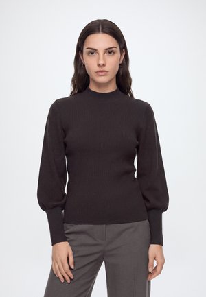 Ung kvinde med langt mørkt hår iført en sort ribstrikket sweater med ballonærmer og grå bukser, står mod en ensfarvet hvid baggrund.