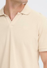 Polo en coton beige avec col en V, manches courtes et logo de marque subtil brodé sur la poitrine. Texture lisse, tissu léger.