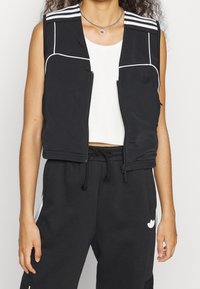 Schwarzes ärmelloses Oberteil mit weißen Abschlussnähten und drei weißen horizontalen Streifen auf den Schultern. Reißverschluss, glatter Stoff und slim Fit.