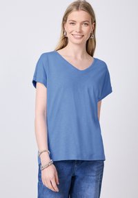 Modrý tílko s krátkým rukávem a výstřihem do V vyrobené z měkkého materiálu. Má volný střih a jemnou texturu. Kombinováno s džíny a náramky.