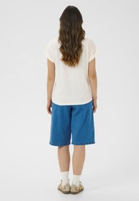 T-shirt en coton crème, coupe décontractée, vue de dos. Associé à un short en denim bleu à mi-cuisse, des chaussettes blanches et des chaussures beiges. Cheveux longs et ondulés.