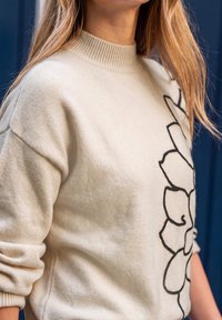 Femme portant un pull crème avec un motif floral abstrait noir sur le côté, debout devant un fond bleu foncé.