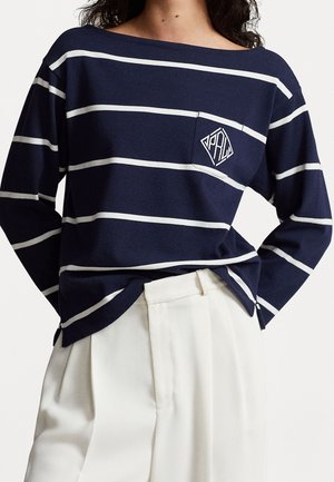 Mujer con camisa azul marino de manga larga con rayas horizontales blancas y pantalones blancos, camisa ligeramente metida en la cintura.