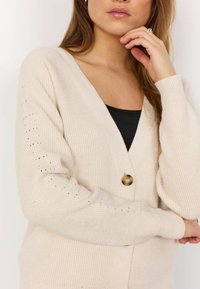 Cardigan bej cu decolteu adânc în V, textură ribbed și mâneci lungi. Prezintă un singur nasture rotund din alamă și detalii mici sub formă de decupaje pe mâneci.