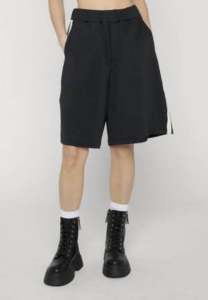 Person iført løstsiddende sorte knælange shorts med hvide sidelommer, hvide sokker og sorte snørebåndskampstøvler, stående mod en ensfarvet baggrund.