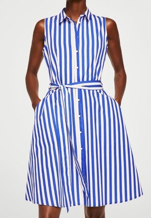 Ärmelloses blau-weiß gestreiftes Kleid mit vertikalen Streifen, Kragen, vorderen Knöpfen, Taillengürtel und Taschen, getragen von einer Person mit den Händen in den Taschen.