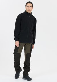 Zwarte fleecejack met een hoge kraag, gecombineerd met donkere olijfgroene cargobroeken met zwarte accenten en meerdere zakken. Draagt donkere schoenen.