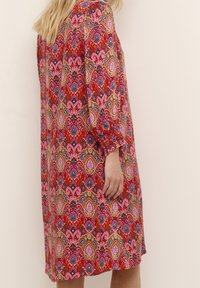 Kvinna i en knälång, långärmad röd klänning med ett flerfärgat paisleymönster som står mot en enfärgad beige bakgrund.