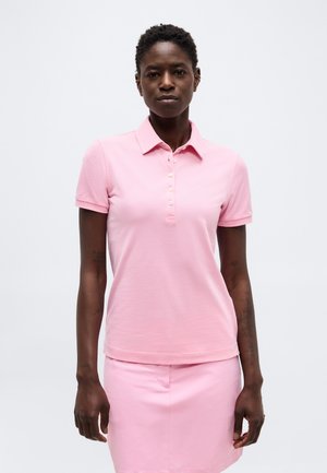 Musta nainen, joka pukeutunut vaaleanpunaiseen lyhythihaiseen poolopaitaan ja vastaavaan hameeseen, seisoo yksivärisen valkoisen taustan edessä.