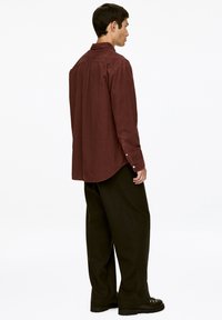 Burgunderfarbenes Cordhemd mit abgerundetem Saum, Knopfmanschetten und Rückenpasse. Kombiniert mit weit geschnittenen schwarzen Hosen und klobigen schwarzen Schuhen.