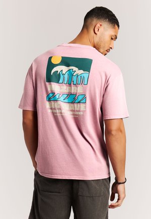 Lichtroze katoenen T-shirt met een grafische print op de achterkant van golven, een zon en de tekst "NAZARÉ BIG WAVE." Korte mouwen, losse pasvorm.