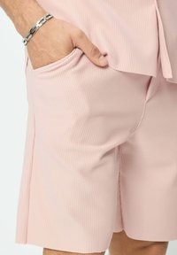 Shorts en tissu côtelé rose clair avec une poche latérale. Vue rapprochée mettant en avant la texture et l'ajustement, accessoirisés d'un bracelet en chaîne argenté.