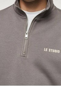 Gray zip-up sweatshirt made of soft fabric featuring a quarter-zip front and white embroidered "LE STUDIO" text on the chest.