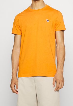 Homme portant un t-shirt manches courtes orange vif avec un petit logo zèbre sur la poitrine, associé à un short beige clair, debout devant un fond uni.