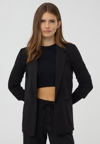 Giovane donna con capelli castano chiaro lisci, indossa un blazer nero sopra un maglione corto e pantaloni neri, in piedi con le mani dietro la schiena.