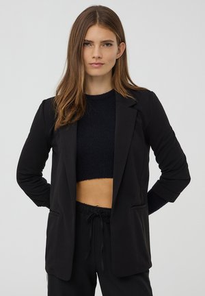 Giovane donna con capelli castano chiaro lisci, indossa un blazer nero sopra un maglione corto e pantaloni neri, in piedi con le mani dietro la schiena.