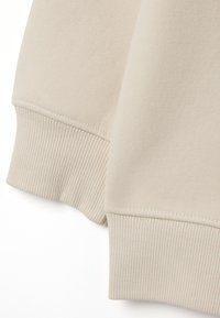 Sudadera beige con puños acanalados y un acabado suave y texturizado. Detalle de costura visible en el dobladillo y los bordes.