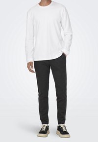 Camicia bianca in cotone a maniche lunghe abbinata a pantaloni grigi scuri e sneakers nere con suola chiara spessa. Texture liscia, vestibilità casual.