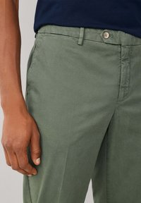 Chinos en coton vert avec une finition texturée, dotés d'une fermeture par bouton et de passants pour ceinture. Une main repose sur le côté du pantalon.
