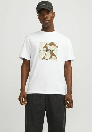 Hvid bomulds t-shirt med et centralt grafisk motiv af brune blade og teksten "Originals Studio". Modellen bærer mørke bukser og en sort kasket.