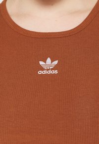 Brązowy prążkowany top na ramiączkach z okrągłym dekoltem, ozdobiony białym haftowanym logo Adidas z trzema paskami i motywem liścia.