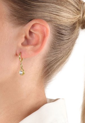 Orecchino a cerchio d'oro con un pendente a forma di goccia che tiene una gemma trasparente su una donna con capelli biondi raccolti.