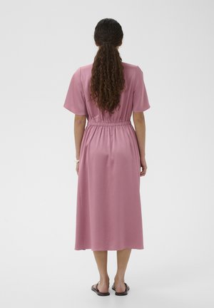 Kvinde med langt krøllet hår iført en pink midi-kjole med korte ærmer og sorte sandaler, stående med ryggen til mod en ensfarvet baggrund.