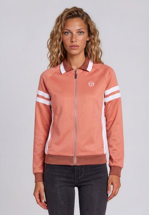 Jeune femme aux cheveux bouclés portant une veste de sport zippée rose et blanche avec des rayures sur les manches, debout devant un fond uni.