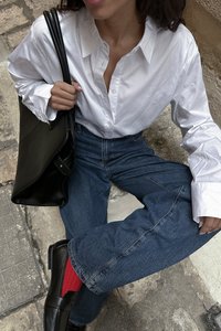 Weißes Hemd mit Knöpfen, dunkelblaue Jeans, schwarze Lederschuhe und rote Socken. Schwarze Ledertasche in der Hand gehalten, sitzend vor einem strukturierten Hintergrund.