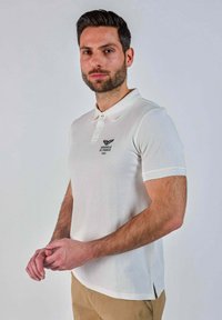 Homme avec des cheveux courts et foncés, portant une barbe et un polo blanc avec le logo "Patrouille de France", se tenant contre un fond clair et uni.