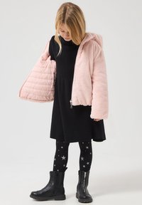 Giacca in pelliccia sintetica rosa chiaro con cappuccio, vestito nero lavorato a maglia, leggings neri con motivo a stelle e stivaletti neri con zip alla caviglia indossati da un bambino.