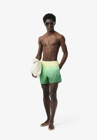 Costumi da bagno con gradiente verde-giallo e vita elasticizzata, abbinati a una borsa tote beige chiaro. La figura indossa occhiali da sole e è scalza.