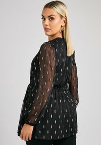 Blusa nera trasparente con maniche lunghe, vita elastica e motivo a foglia metallica. Tessuto morbido con un design vaporoso e dettagli di leggero arricciamento.