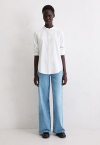 Biała koszula na guziki w pionowe paski z kołnierzem typu mandaryński, zestawiona z jasnoniebieskimi szerokimi spodniami jeansowymi i czarnymi butami na platformie.