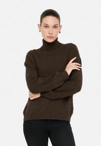 Vrouw met een donkerbruin gespikkelde coltrui en zwarte broek, met gekruiste armen, draagt gouden hartvormige oorbellen, neutrale achtergrond.