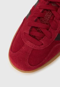 Piros velúr sportcipő fekete díszítésekkel, texturált részletekkel és piros cipőfűzővel. Lekerekített orral és gumitalpú talppal rendelkezik a jobb tapadás érdekében.