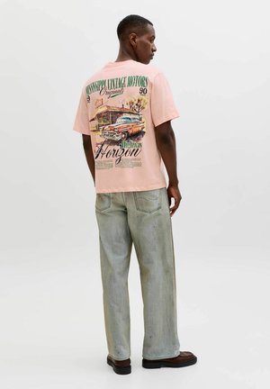 Mężczyzna w jasnoróżowej koszulce z grafiką vintage samochodu i stacji benzynowej, jasnych, wyblakłych jeansach oraz ciemnych butach, stoi na białym tle.