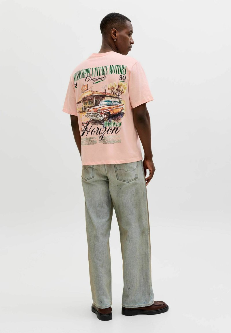 Mann trägt ein hellrosa T-Shirt mit Vintage-Auto- und Tankstellen-Grafik, hell gewaschene Jeans und dunkle Schuhe, steht vor weißem Hintergrund.