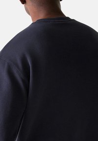 Persona vistiendo una sudadera lisa de color azul marino oscuro, vista desde atrás sobre un fondo liso de color gris claro.