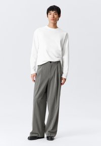 Une jeune personne portant une chemise blanche à manches longues, un pantalon large à rayures grises et des chaussures noires, sur un fond clair uni.