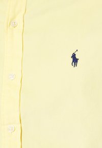 Žlutá košile na knoflíky z žebrované tkaniny s tříknoflíkovou légou. Na levé straně hrudi je malá námořnicky modrá výšivka hráče póla.